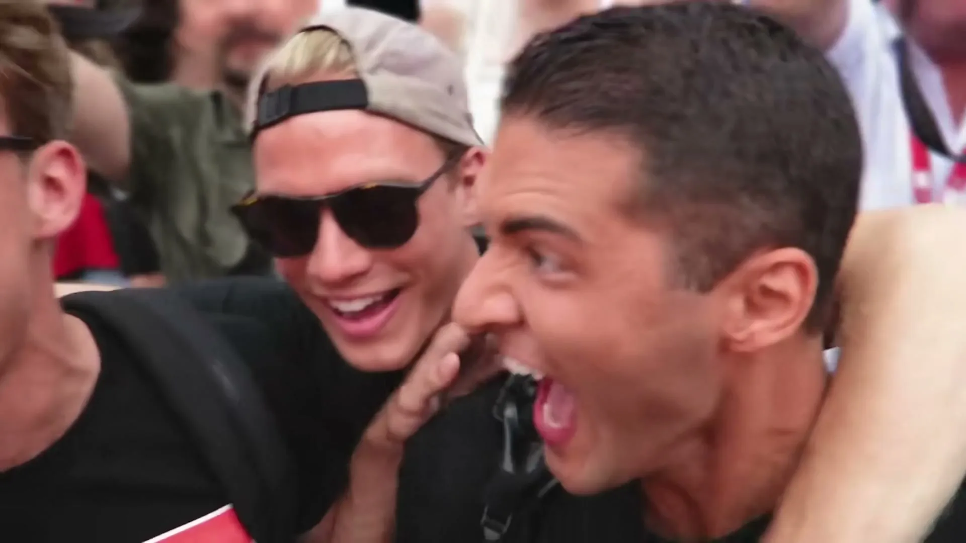 Two young men, one with a backwards cap and sunglasses, the other with dark hair, are cheering excitedly in a crowd.