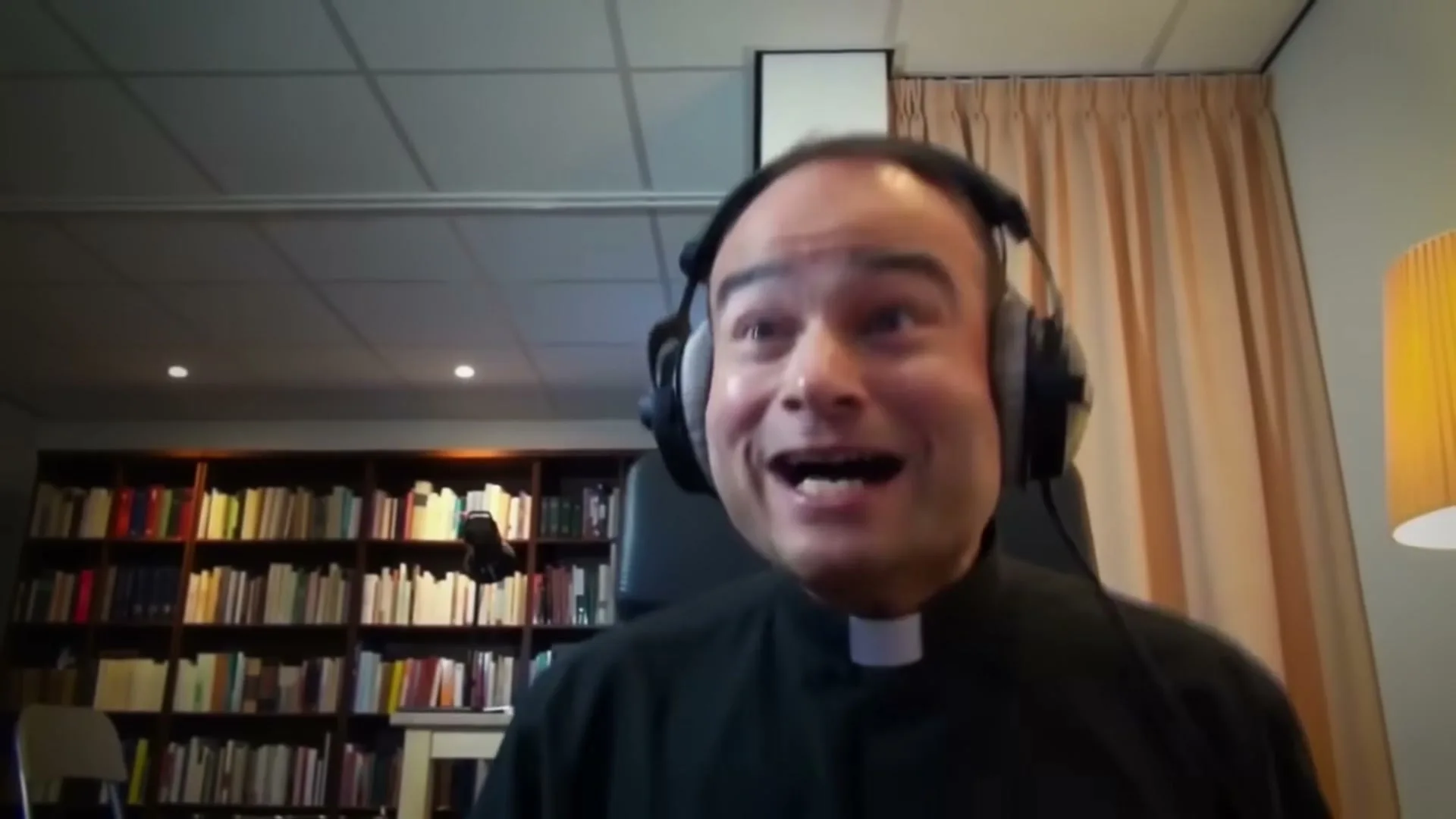 A man in his 30s, wearing a priest's collar and headphones, laughs joyfully while looking at a screen in a room with bookshelves.