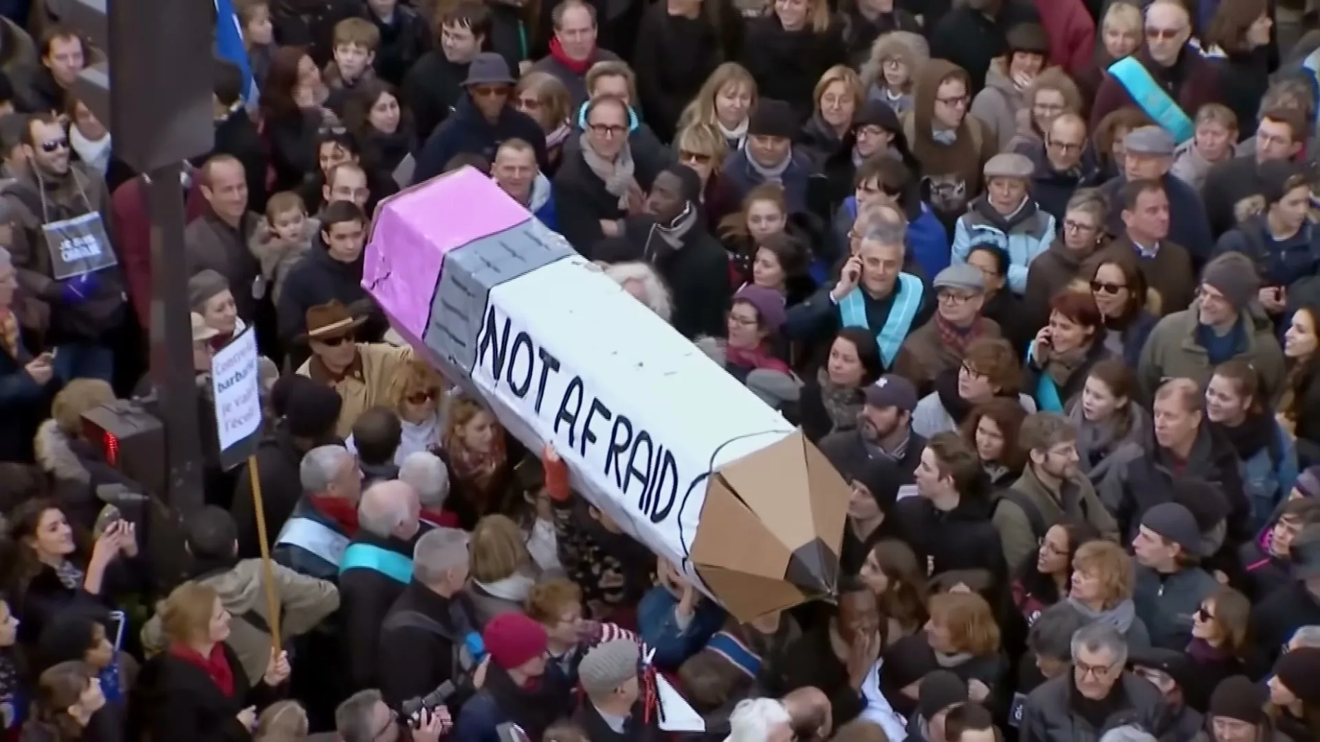 A large crowd of diverse people are gathered outdoors, holding up a giant pencil prop that reads "NOT AFRAID" in black letters.