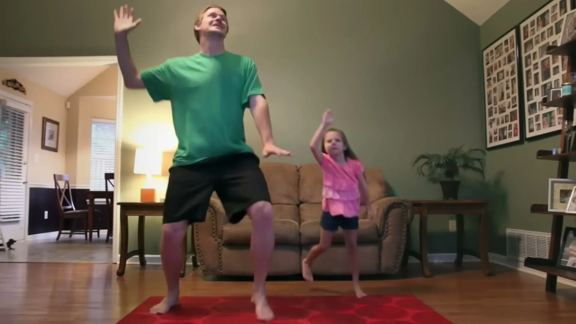 A father and his young daughter dance barefoot on a red rug in their living room, both smiling and moving their arms.