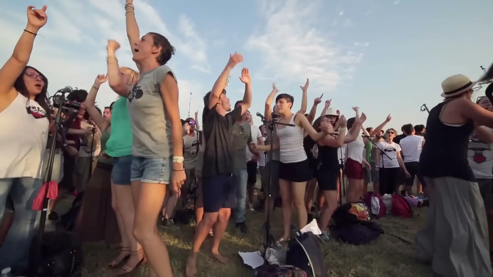A diverse group of young adults, some barefoot, joyfully sing and dance with their arms raised in an outdoor field under a bright sky.