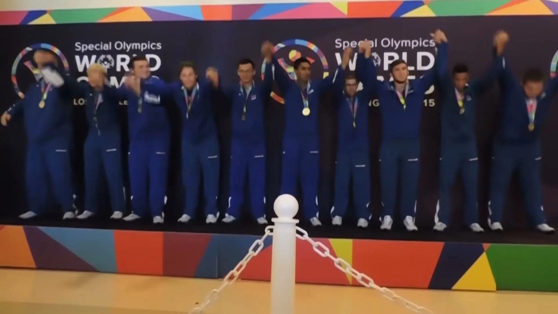 A group of male athletes in blue tracksuits with medals around their necks stand on a podium, raising their arms in triumph at the Special Olympics World Games.