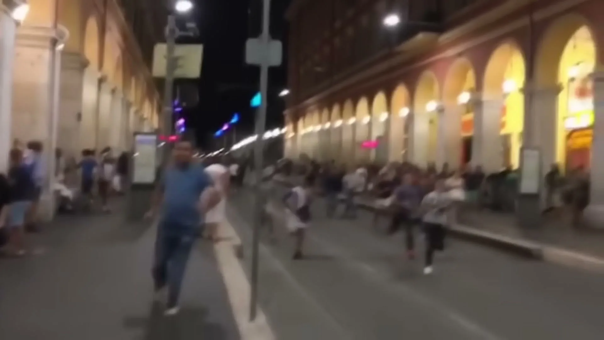 A chaotic scene unfolds at night on a city street as a crowd of people, including a man in a blue shirt, run away from something unseen.