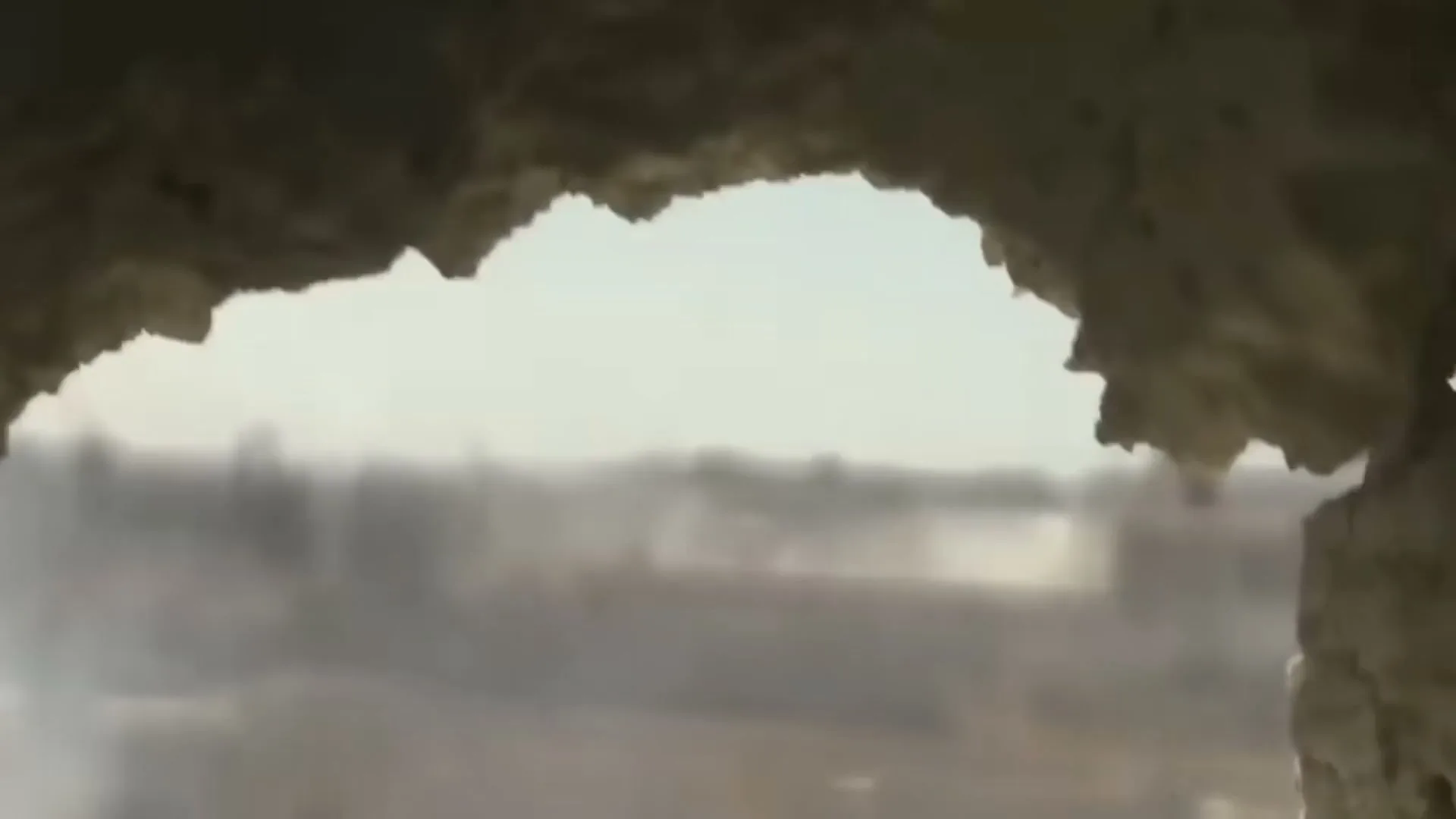 A wide shot through a broken wall shows a war-torn city with smoke rising from buildings under a hazy sky.