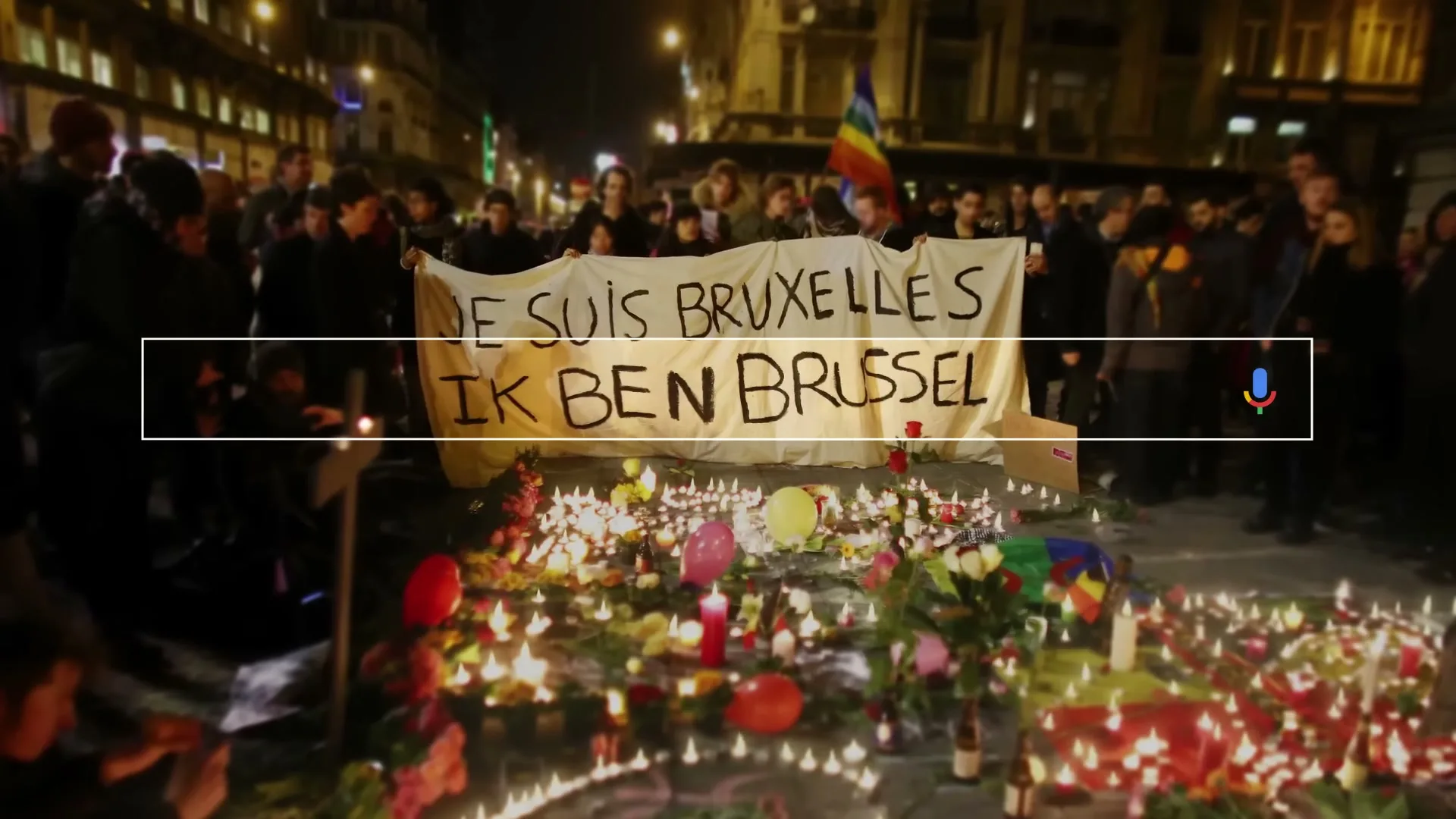 A crowd of people hold a banner that reads "JE SUIS BRUXELLES IK BEN BRUSSEL" above a memorial of candles and flowers at night.