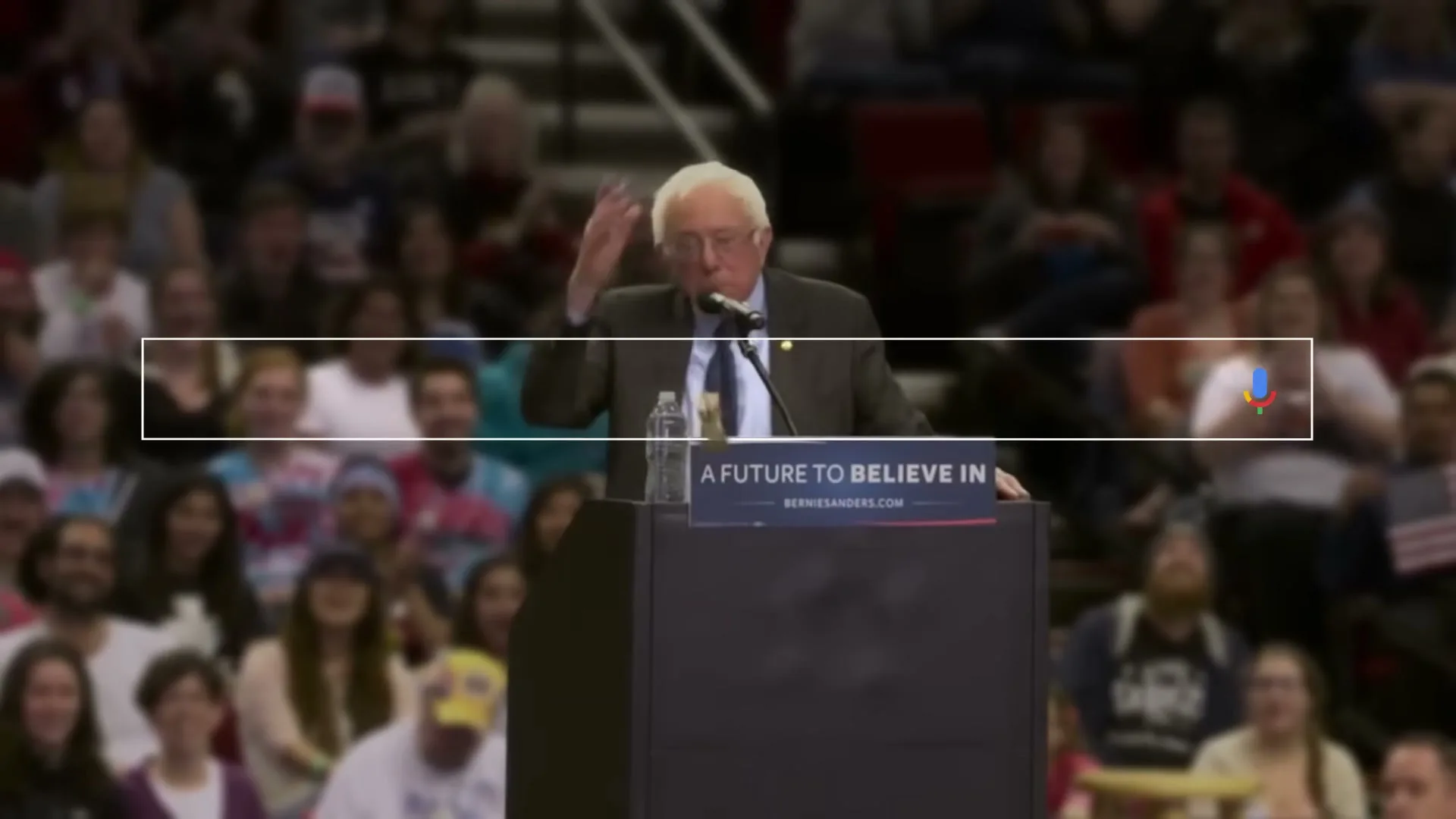 Bernie Sanders, an older white man in a suit, speaks at a podium to a large crowd in an arena, with a Google search bar overlay.