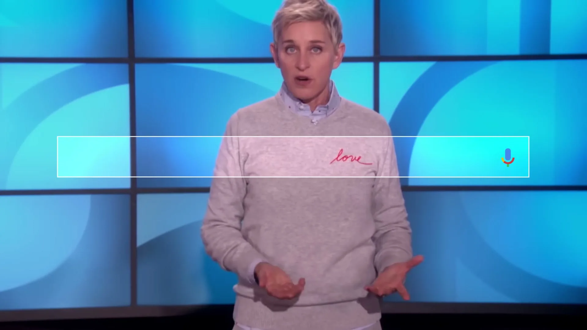 Ellen DeGeneres, a blonde woman in a grey sweater, stands on a stage with a blue grid background, speaking with open hands.