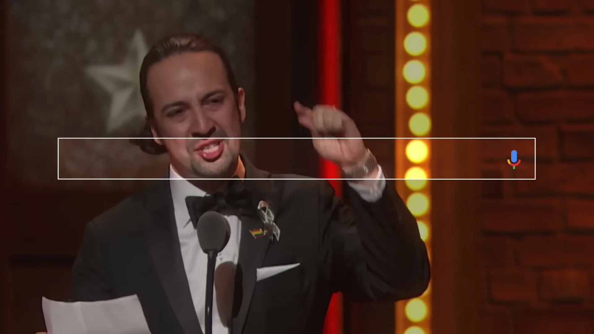 Lin-Manuel Miranda, a male in his 30s, wearing a black tuxedo, speaks passionately into a microphone while holding papers at an awards ceremony.