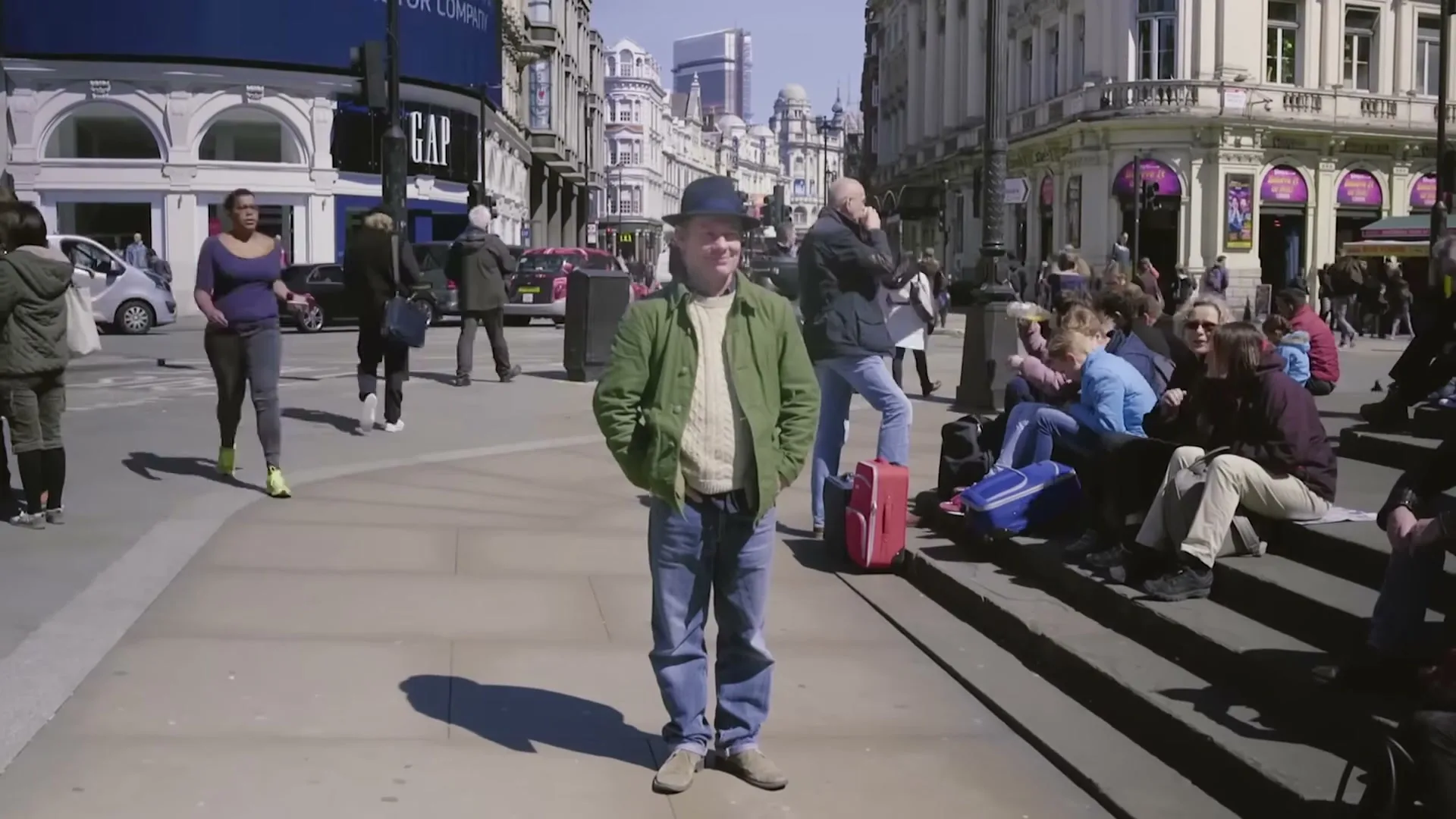 A man in a green jacket and hat stands in the middle of a busy city street, looking directly at the camera.
