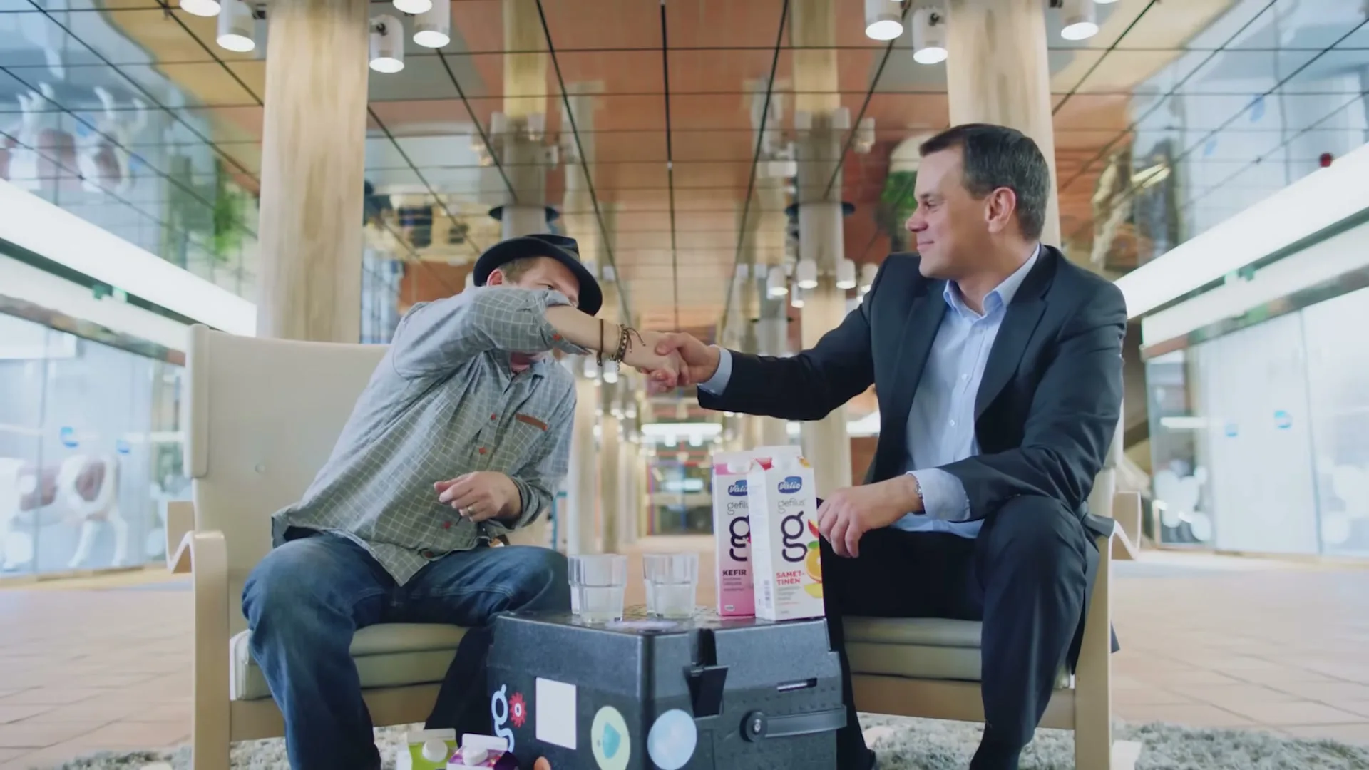 Two men, one in a suit and one in casual clothes, shake hands in a modern, brightly lit interior space with reflective ceilings.