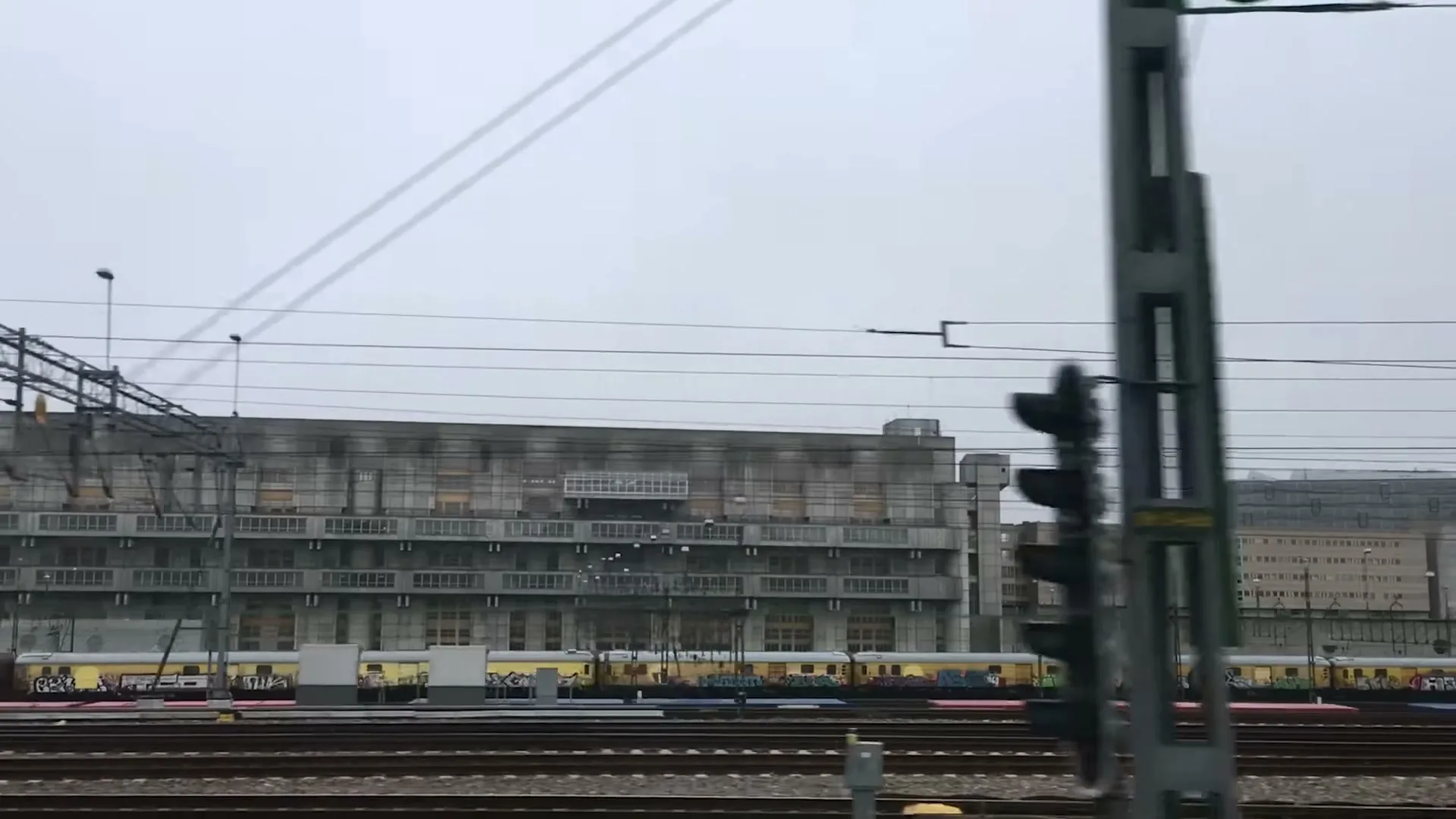 A wide shot from a moving train shows a large, grey industrial building with many windows, several train tracks, and yellow trains covered in graffiti under a grey sky.