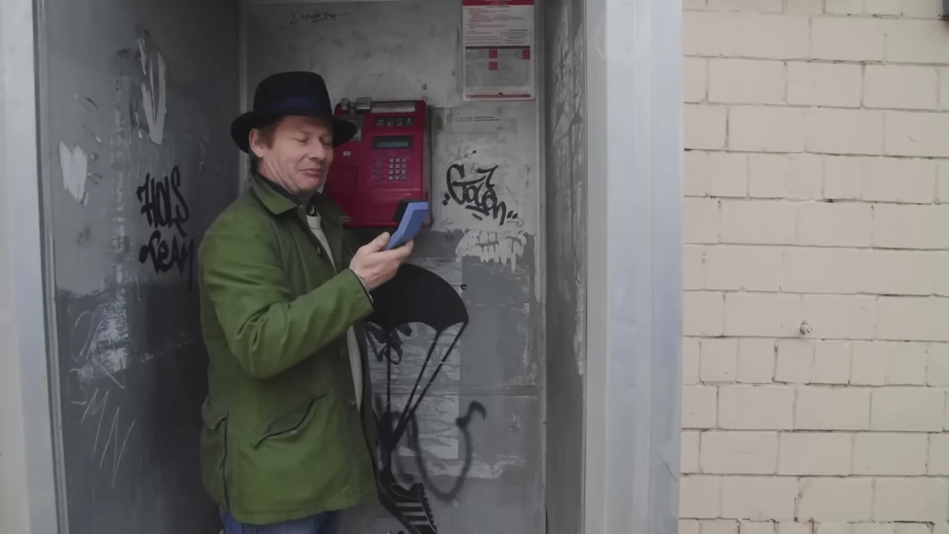 A man in a green jacket and black hat licks a blue phone receiver in a graffiti-covered phone booth.