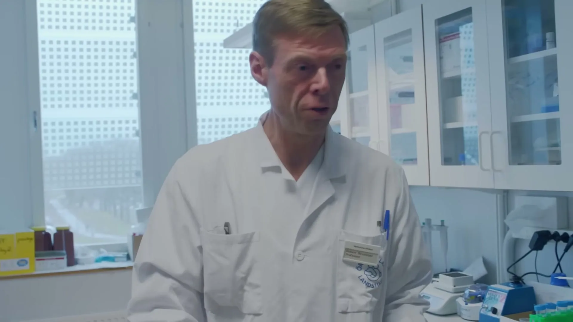 A male scientist in a lab coat looks down, then up, in a bright laboratory with large windows.