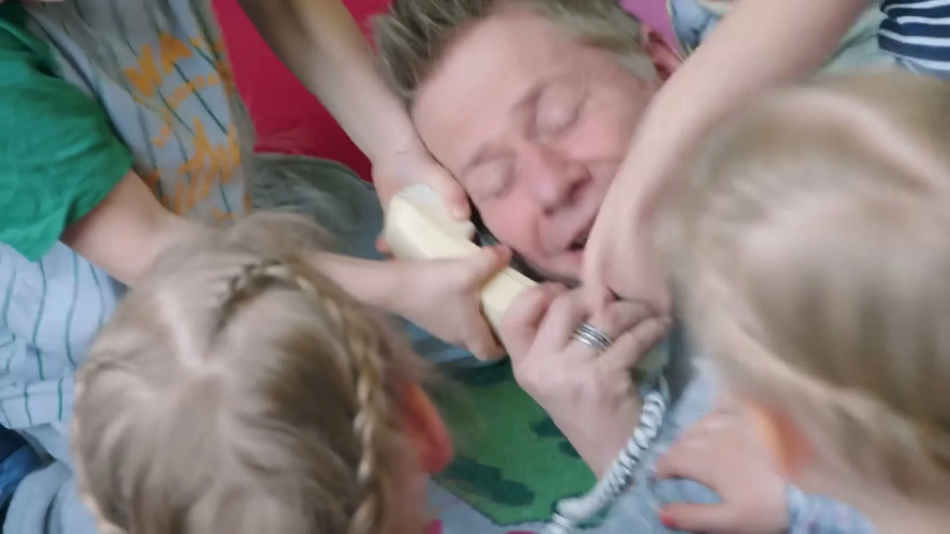 A man in a blue shirt is overwhelmed by several young children climbing on him and grabbing a phone, in a playful, chaotic scene indoors.
