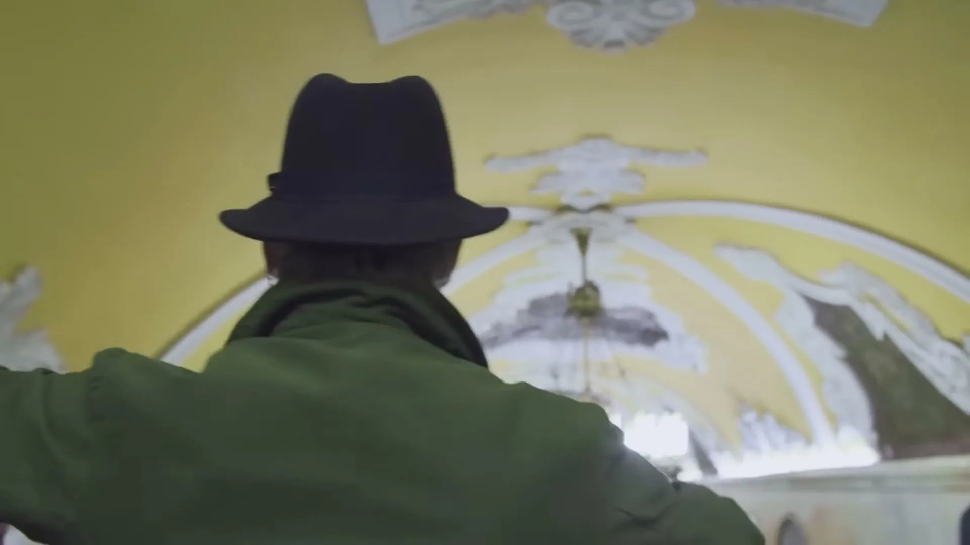 A man in a hat and green coat turns to face the camera, then turns away to walk down a crowded subway station escalator.