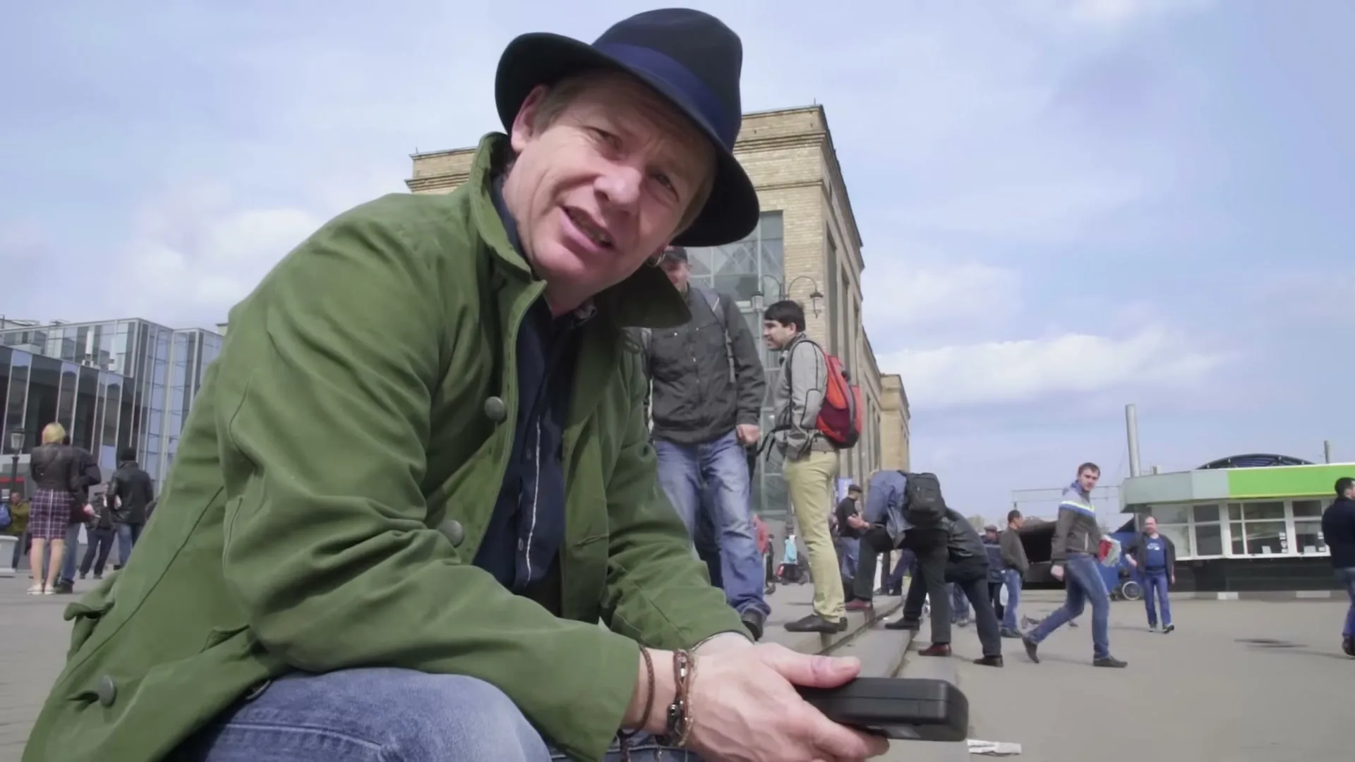 A man in a green coat and black hat sits on the ground, talking to the camera in a busy outdoor plaza with people walking around.