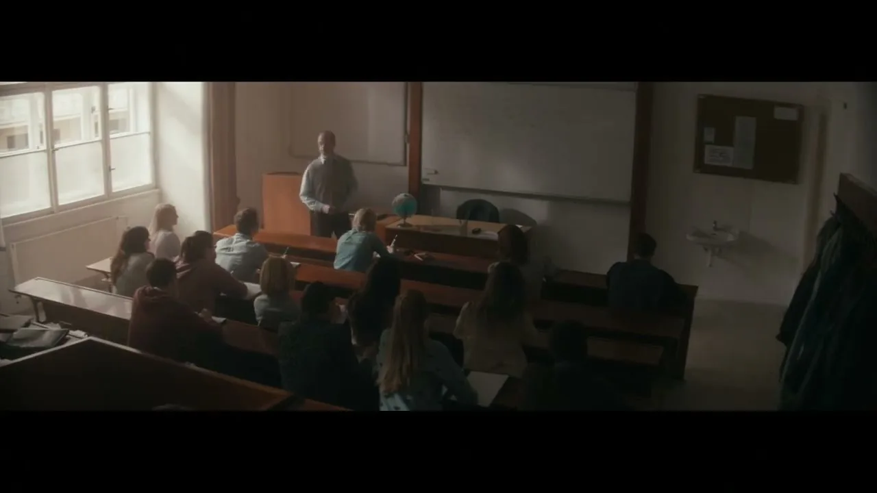 A male teacher stands at the front of a classroom, addressing a group of students seated at wooden desks, with sunlight streaming through a window.