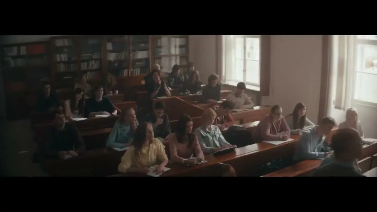 A wide shot shows a classroom full of students sitting at wooden desks, taking notes, with bookshelves in the background.