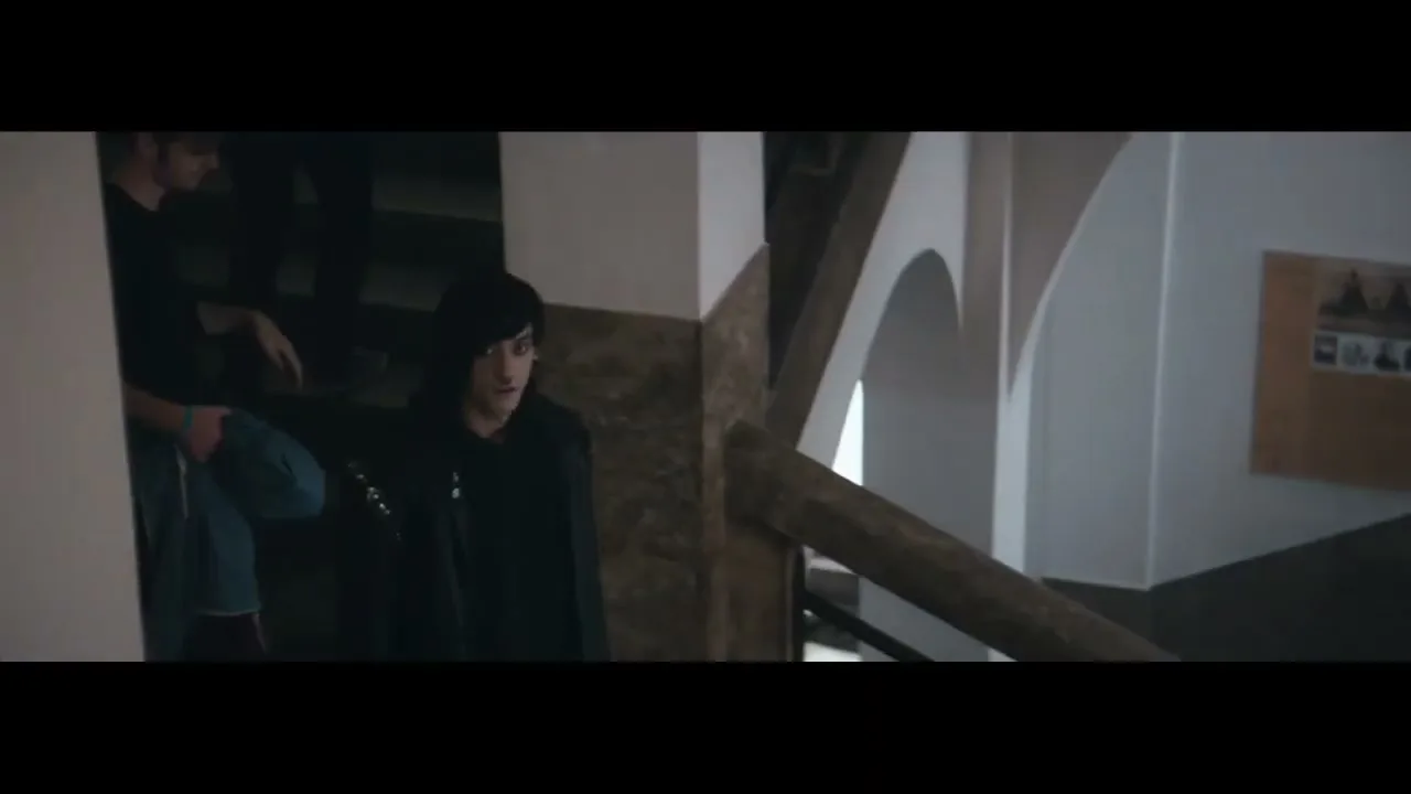 A young man with dark hair and a black hoodie looks around nervously while walking down a staircase in a building.