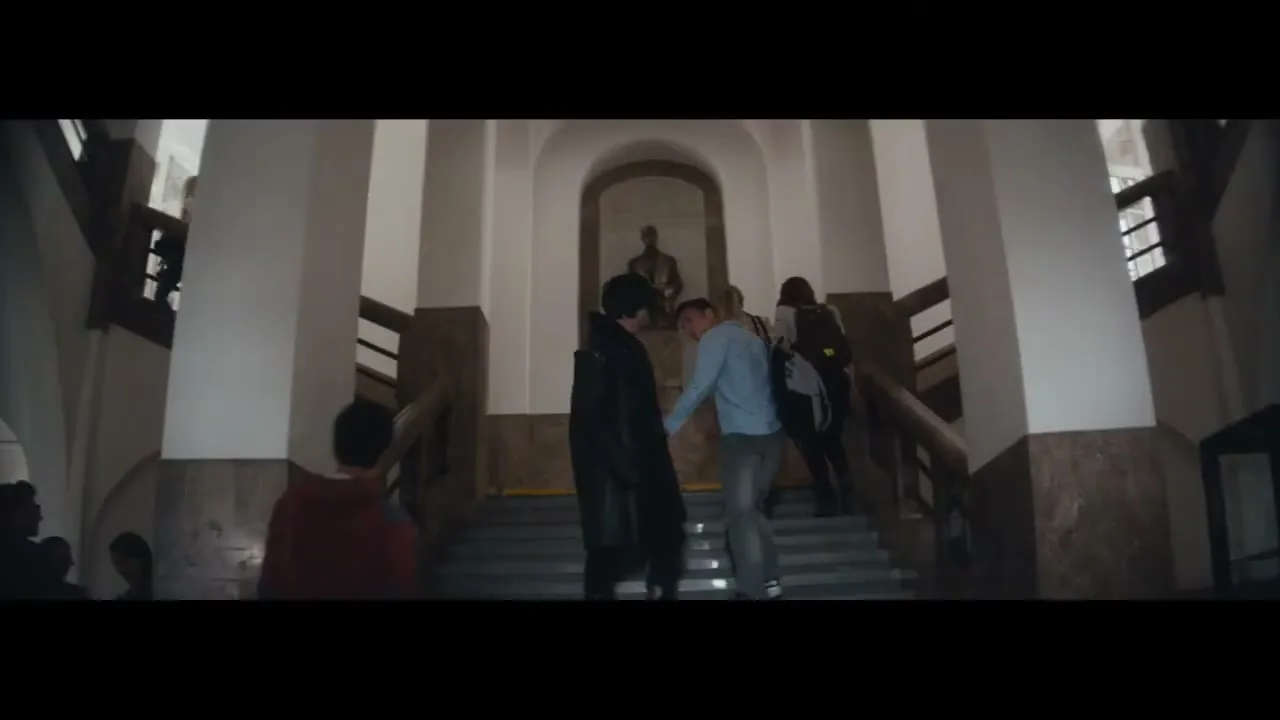 A young man in a long black coat walks up a grand staircase in a school building, followed by other students.