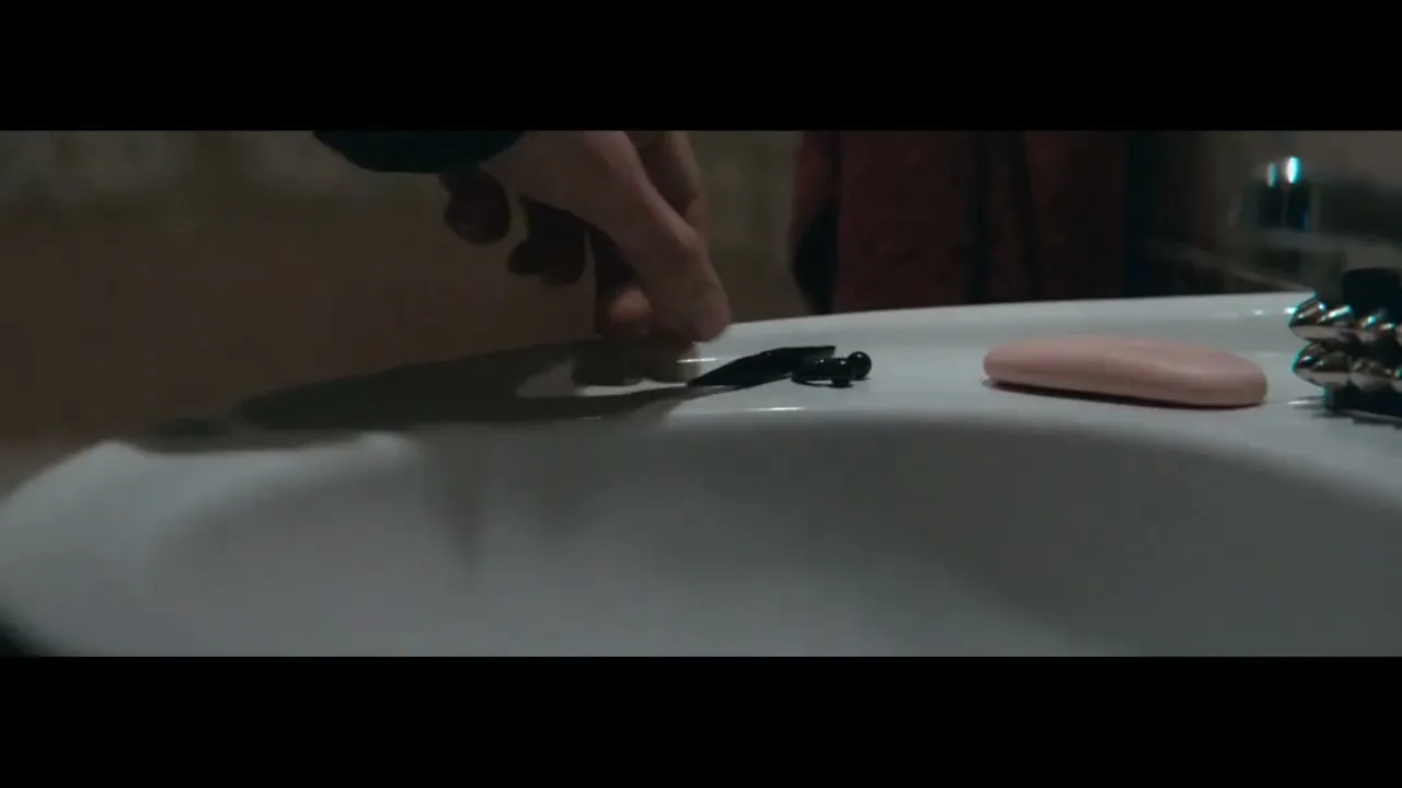A person's hand places a small, black, insect-like object into a white bathroom sink next to a bar of pink soap.