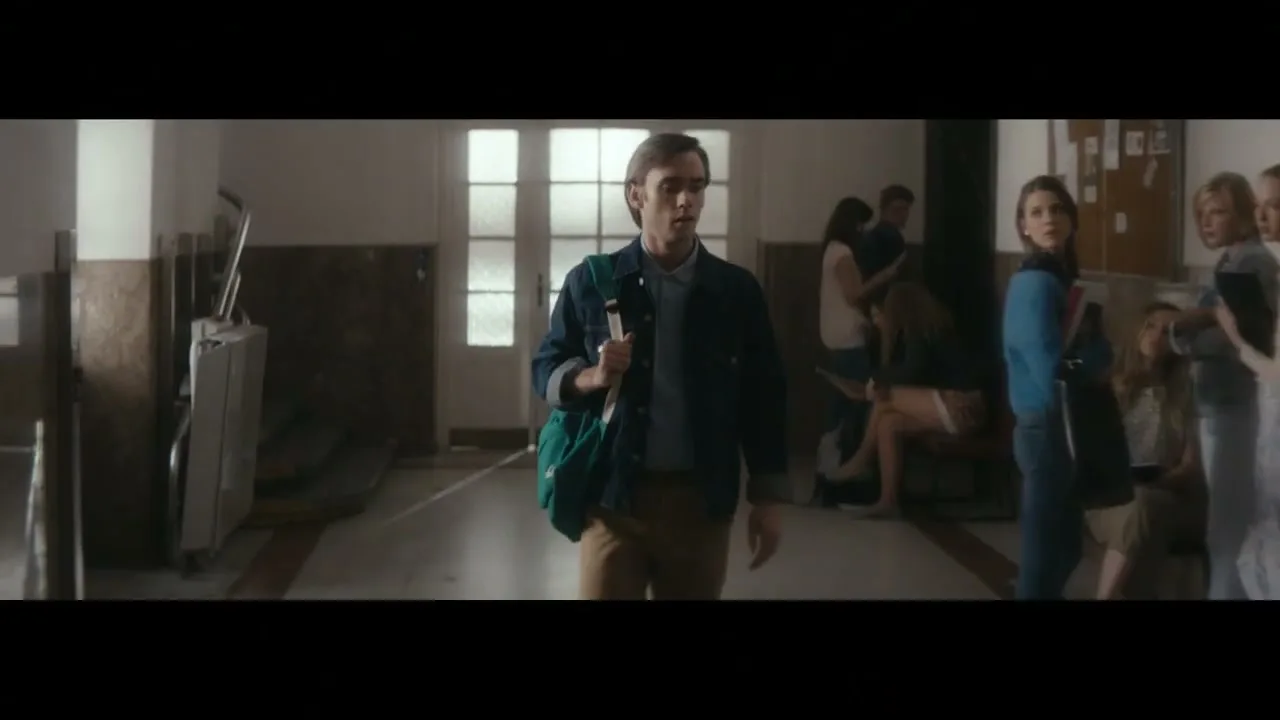 A young man with a green bag walks through a school hallway, looking around as other students gather in the background.