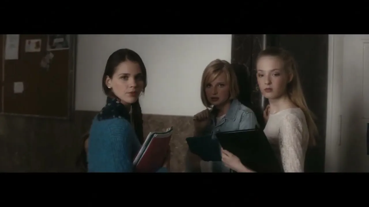 Three teenage girls, two holding folders, stand in a school hallway, looking intently at something off-screen.