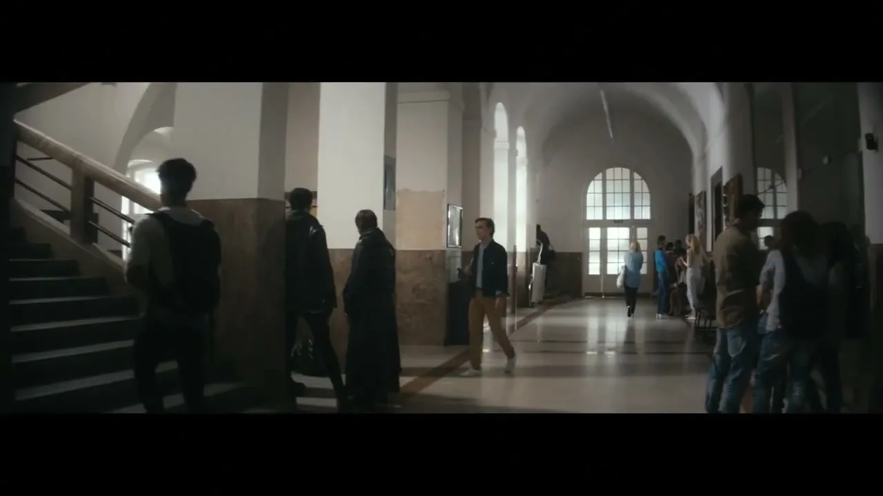 A young man in a blue jacket and khaki pants walks down a bright hallway in a school, passing other students.