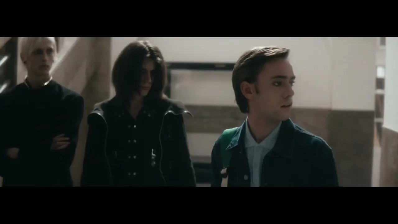 A young man with a backpack looks around nervously while two other people stand behind him in a school hallway.