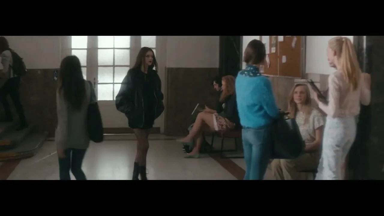 A young woman in a black jacket and fishnet stockings walks through a school hallway, while other students sit and stand around her.