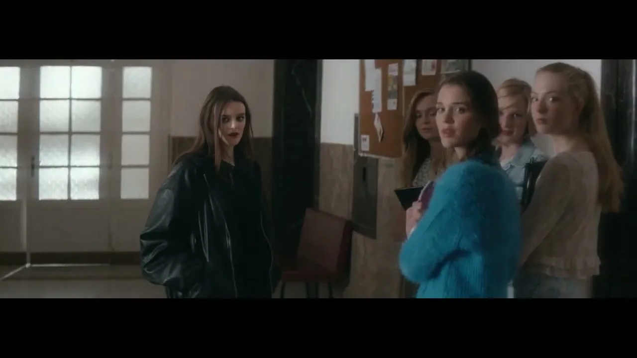 A young woman in a black leather jacket stands opposite a small group of young women in a school hallway, looking at them with a serious expression.