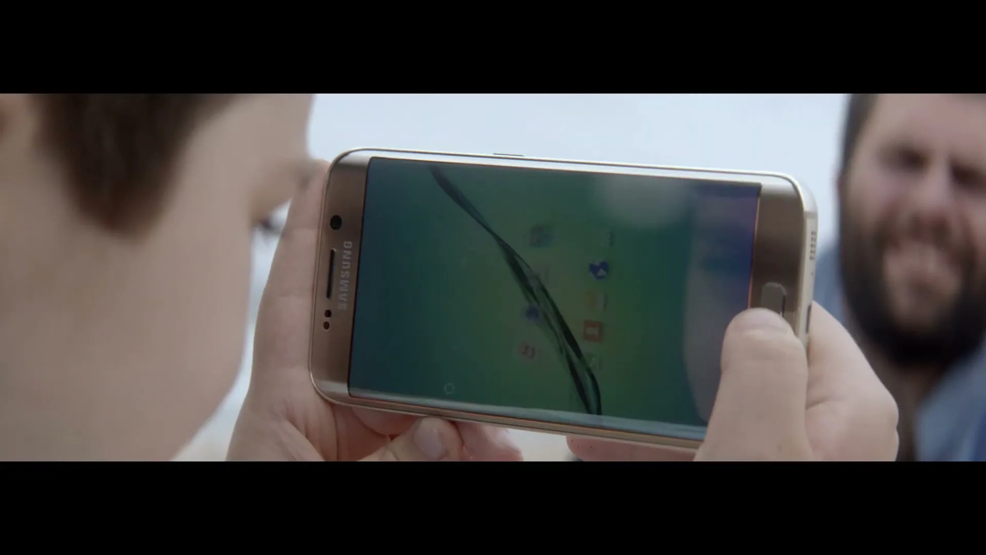A young boy holds a gold Samsung phone horizontally, looking at the screen, while a man with a beard smiles in the blurred background.