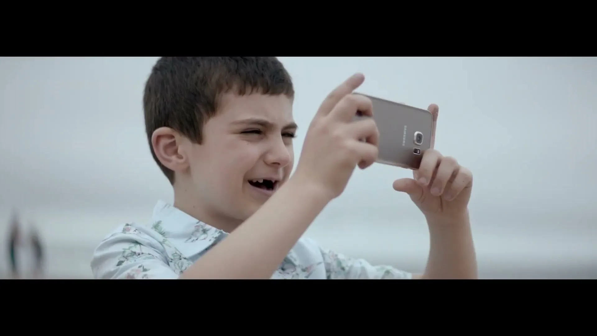 A young boy with a missing front tooth smiles while holding a Samsung phone to take a picture at the beach.