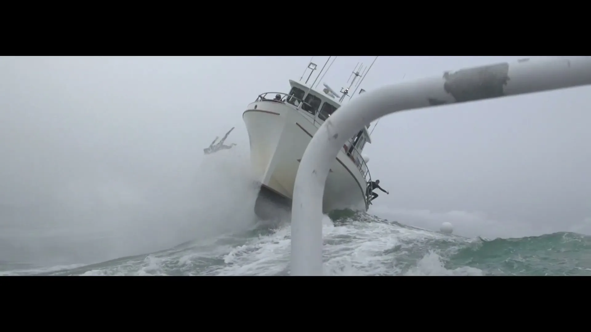 A white boat is seen from a low angle, being tossed by large waves in a stormy, grey ocean, with two people in wetsuits jumping off.
