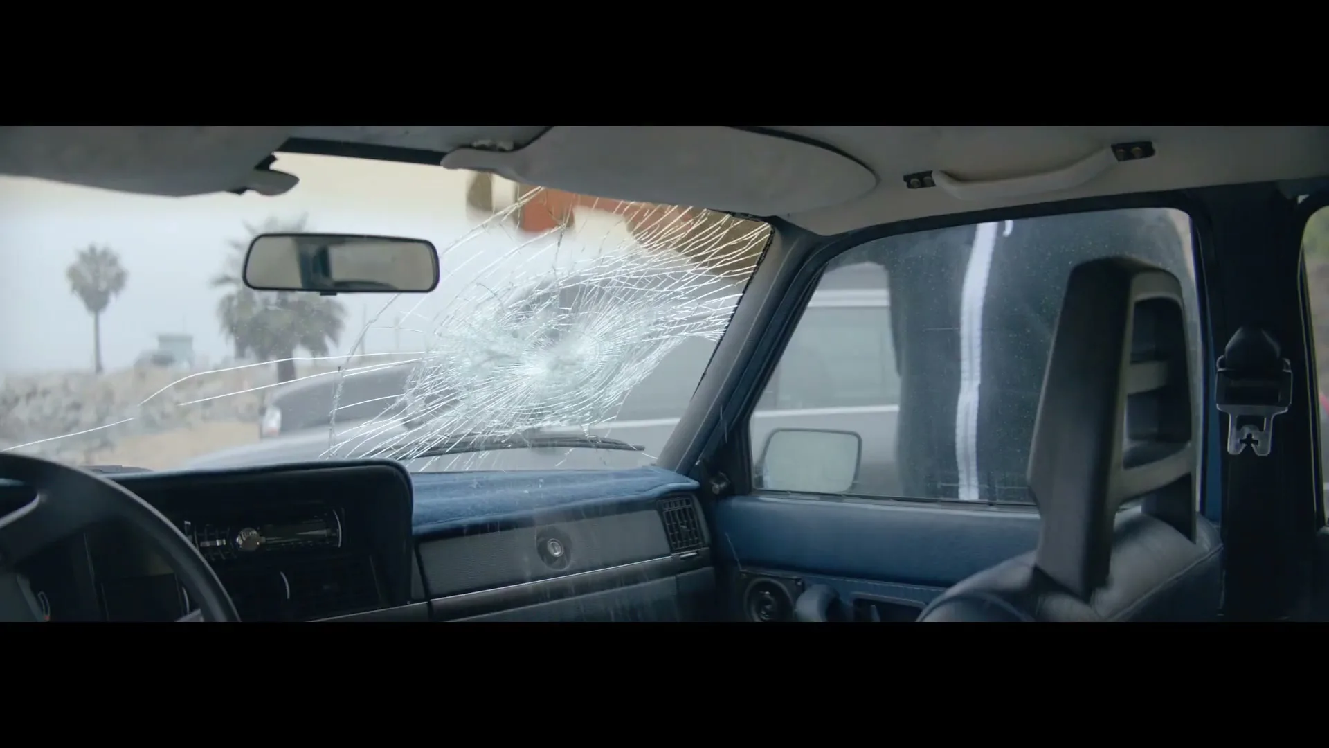 A person in a black and white tracksuit smashes the windshield of a blue car, seen from inside the car on a foggy day near palm trees.