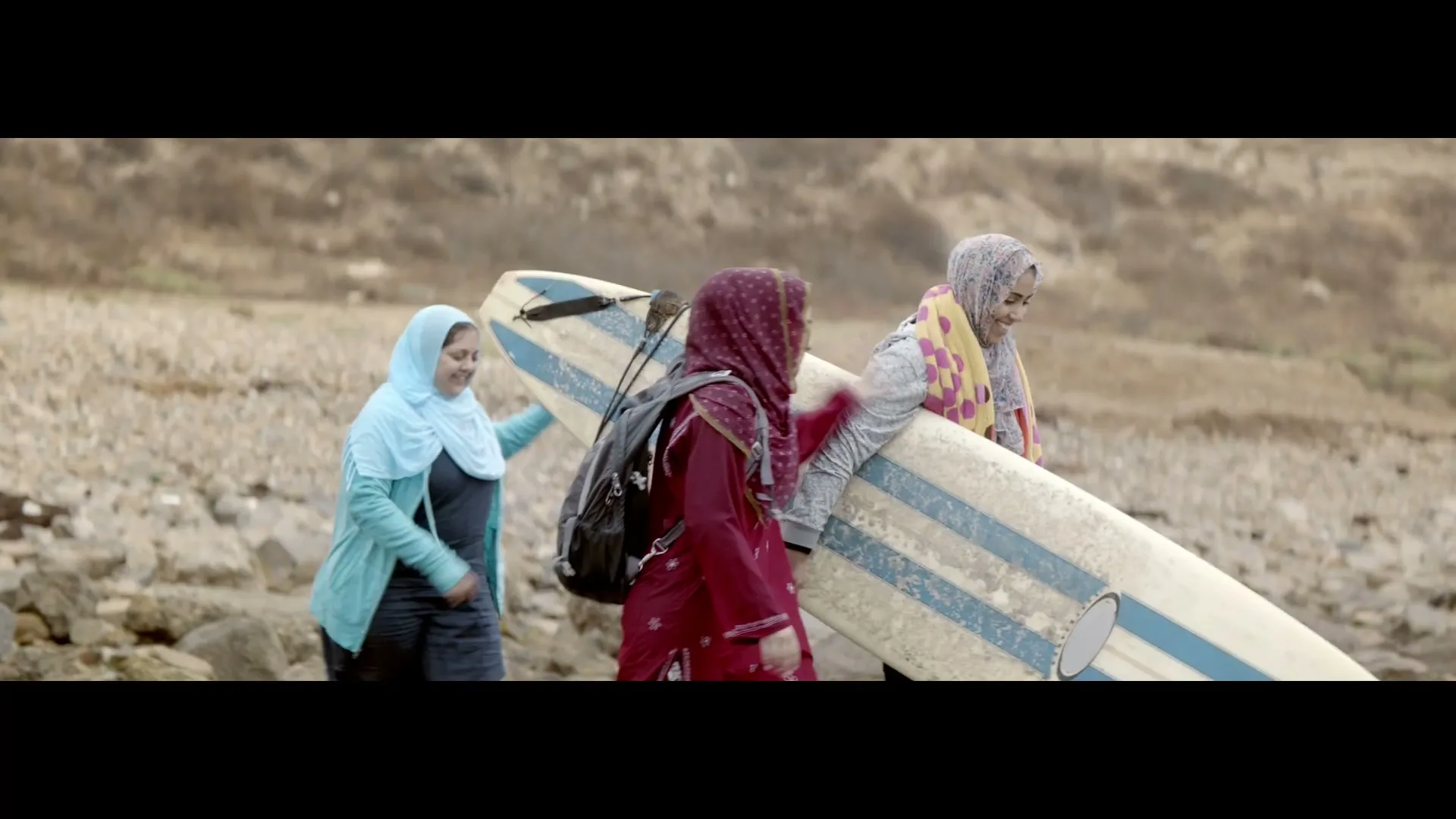 Three young women in hijabs and modest clothing carry a surfboard across a rocky beach, smiling and walking towards the right side of the frame.