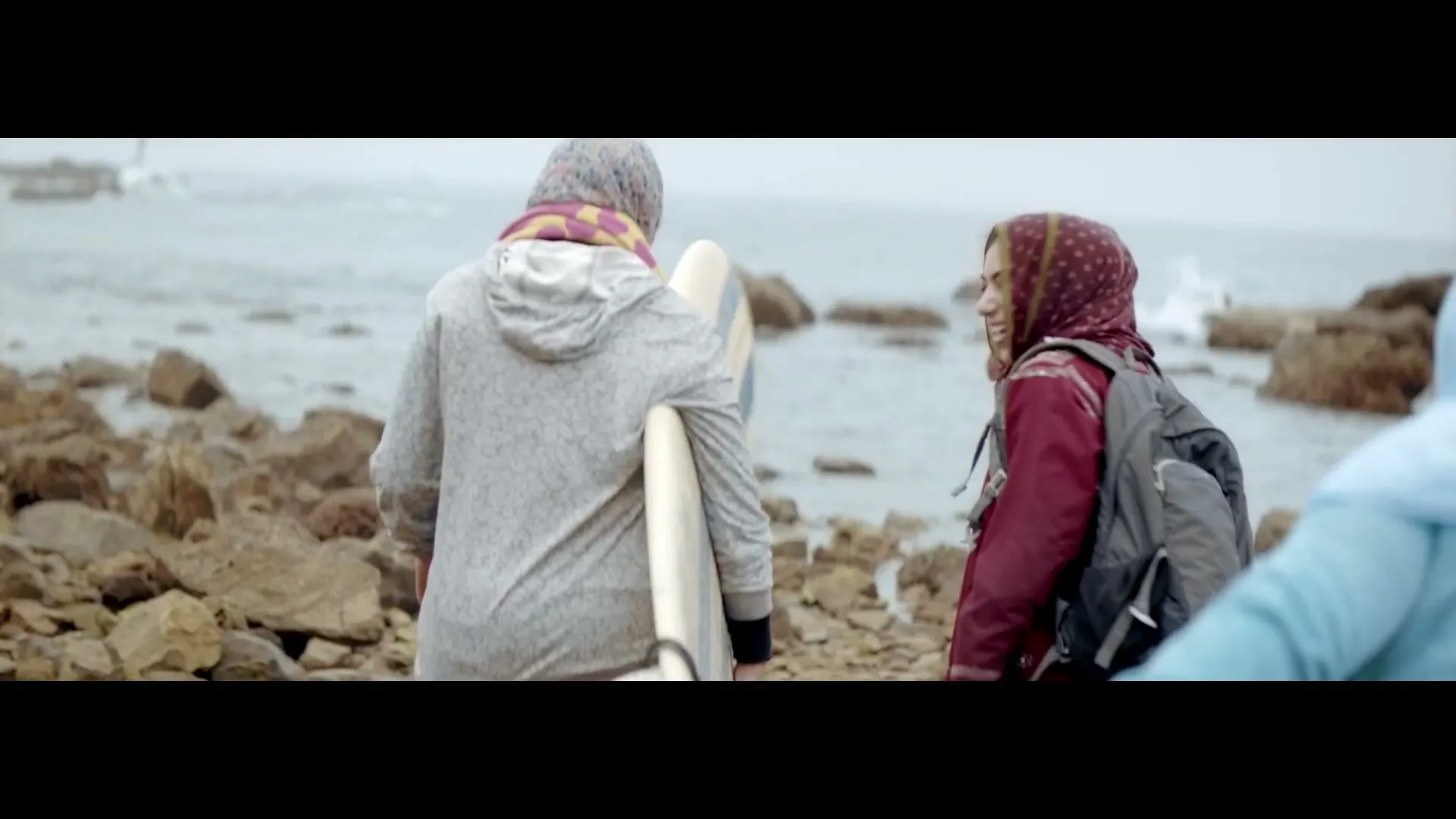 Three young women, two wearing hijabs, walk along a rocky beach with a surfboard, one turning to smile and laugh.