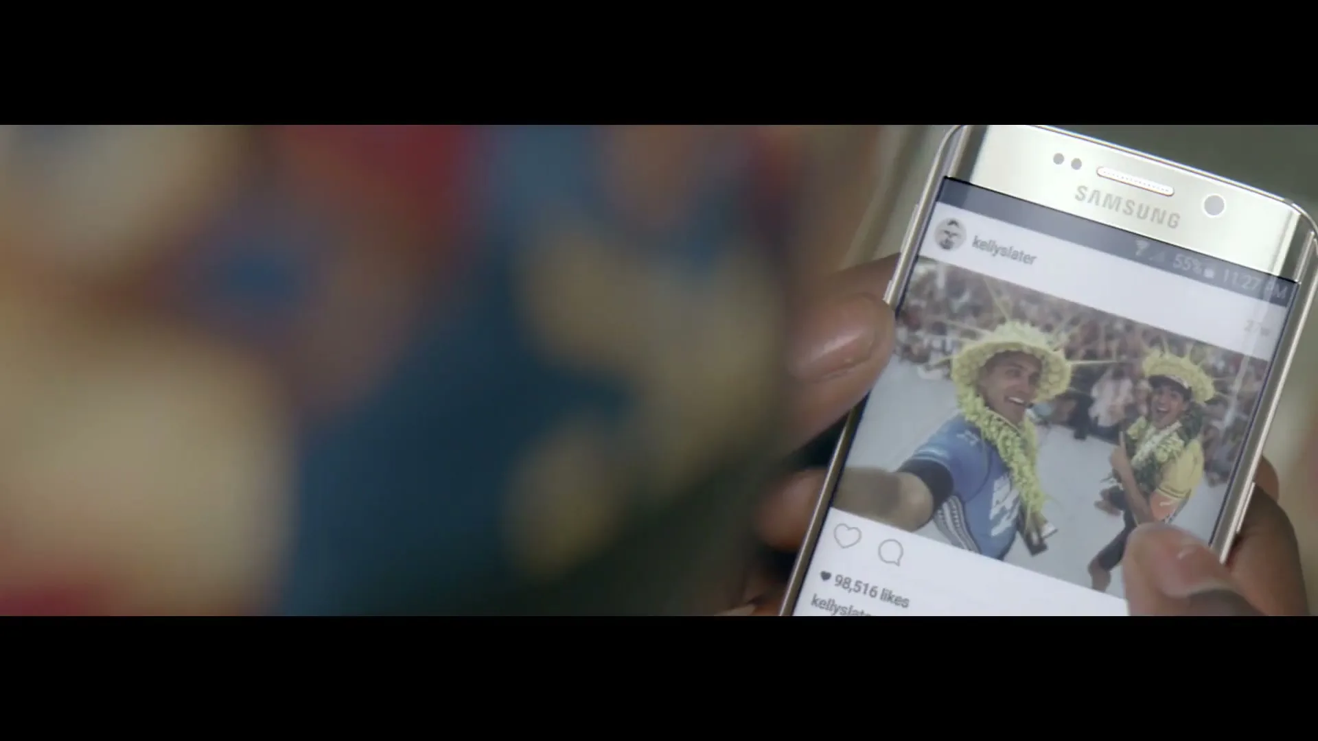 A person holds a Samsung phone displaying an Instagram post of two surfers wearing straw hats and leis, smiling at a crowded event.