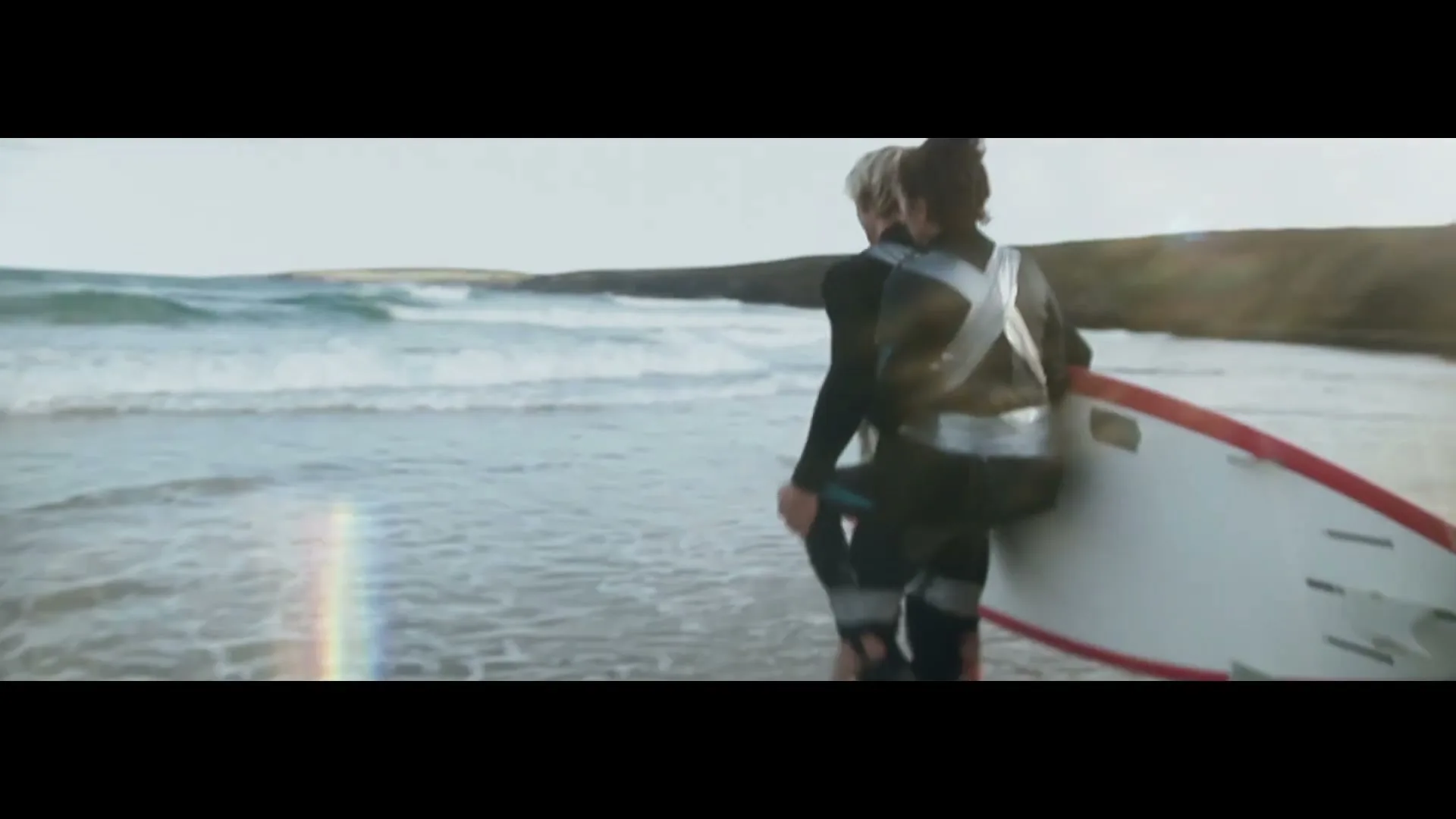 Two surfers, one with silver tape on their back, walk into the ocean with a surfboard on a sunny day.