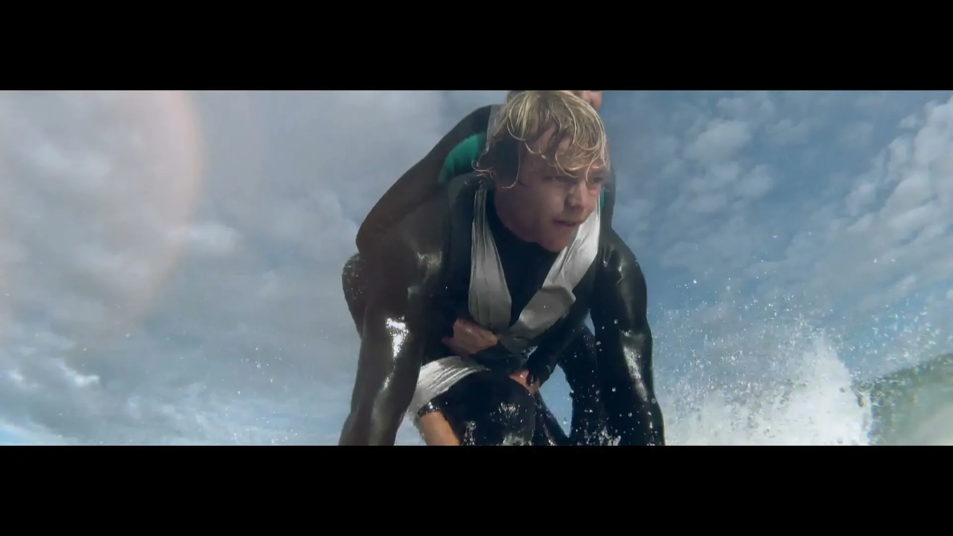 A male surfer with blonde hair in a black wetsuit rides a wave, with another person on his back, under a cloudy sky.