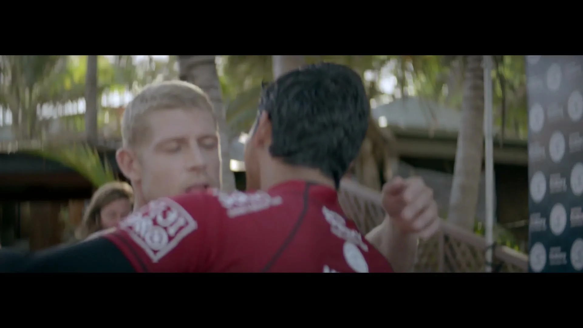 Two male surfers, one with blonde hair and one with dark hair, hug each other outdoors near palm trees and a sponsor banner.