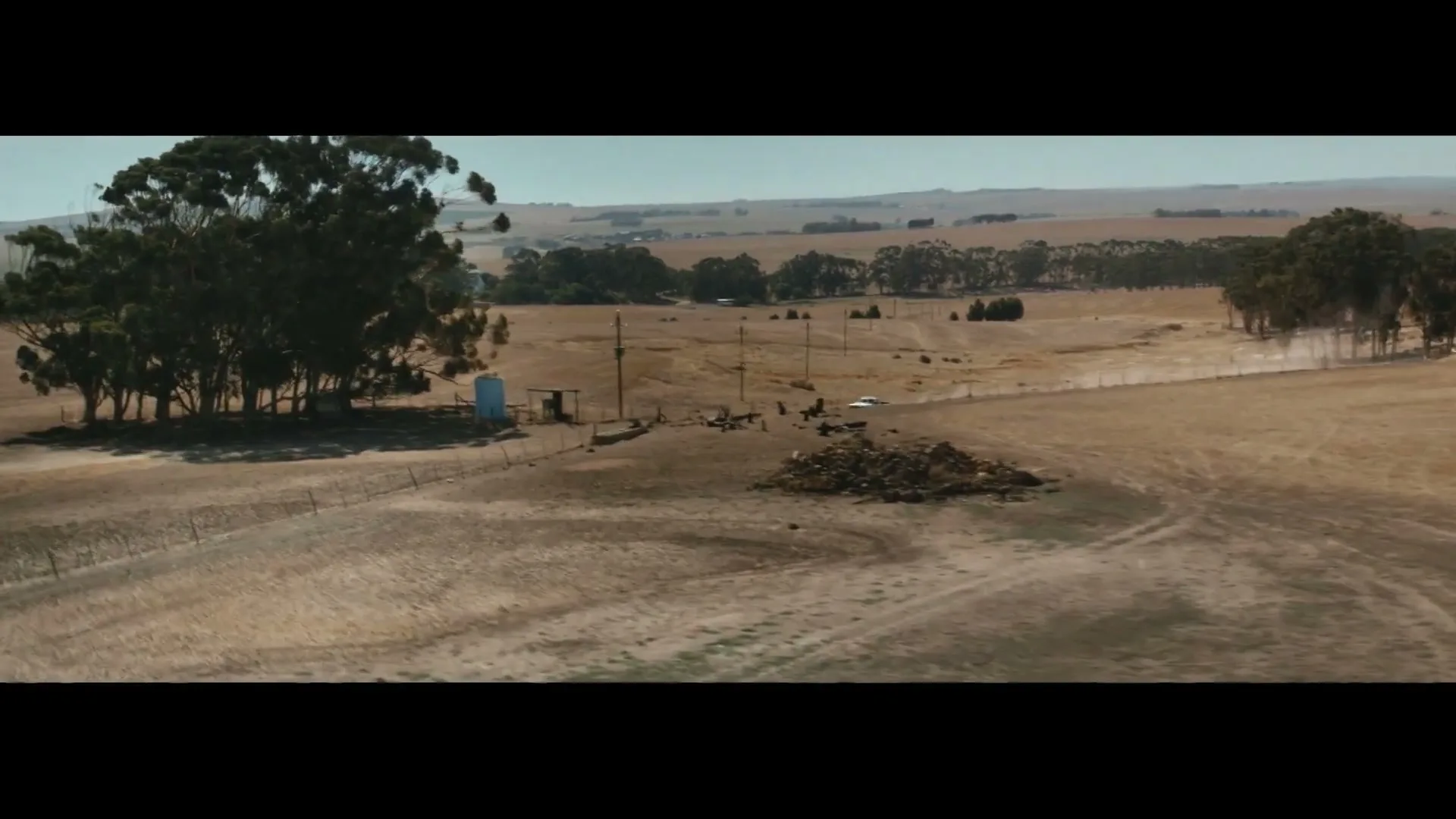A wide shot shows a white car driving on a dirt road through a dry, rural landscape with scattered trees under a clear sky.