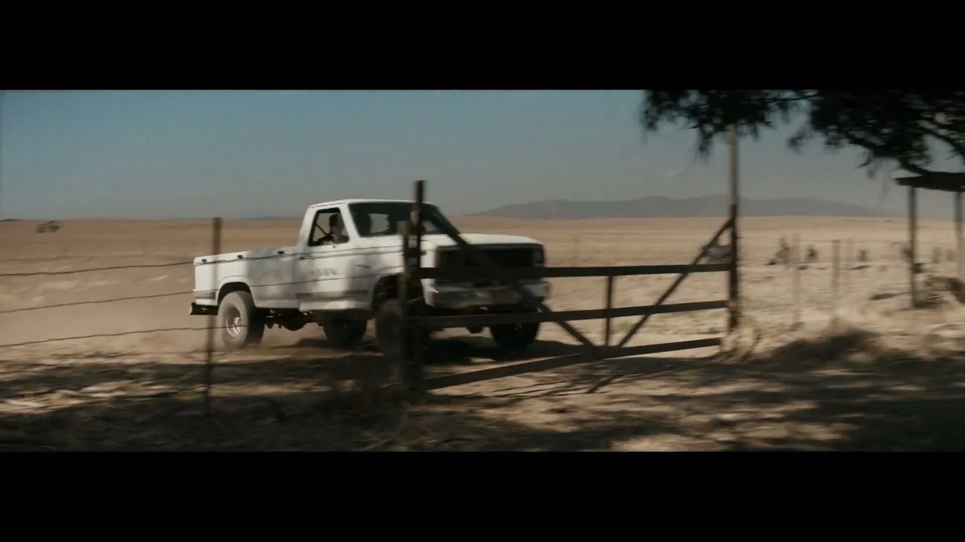 A white pickup truck drives through a dusty, arid landscape, crashing through a wooden fence and kicking up dirt.