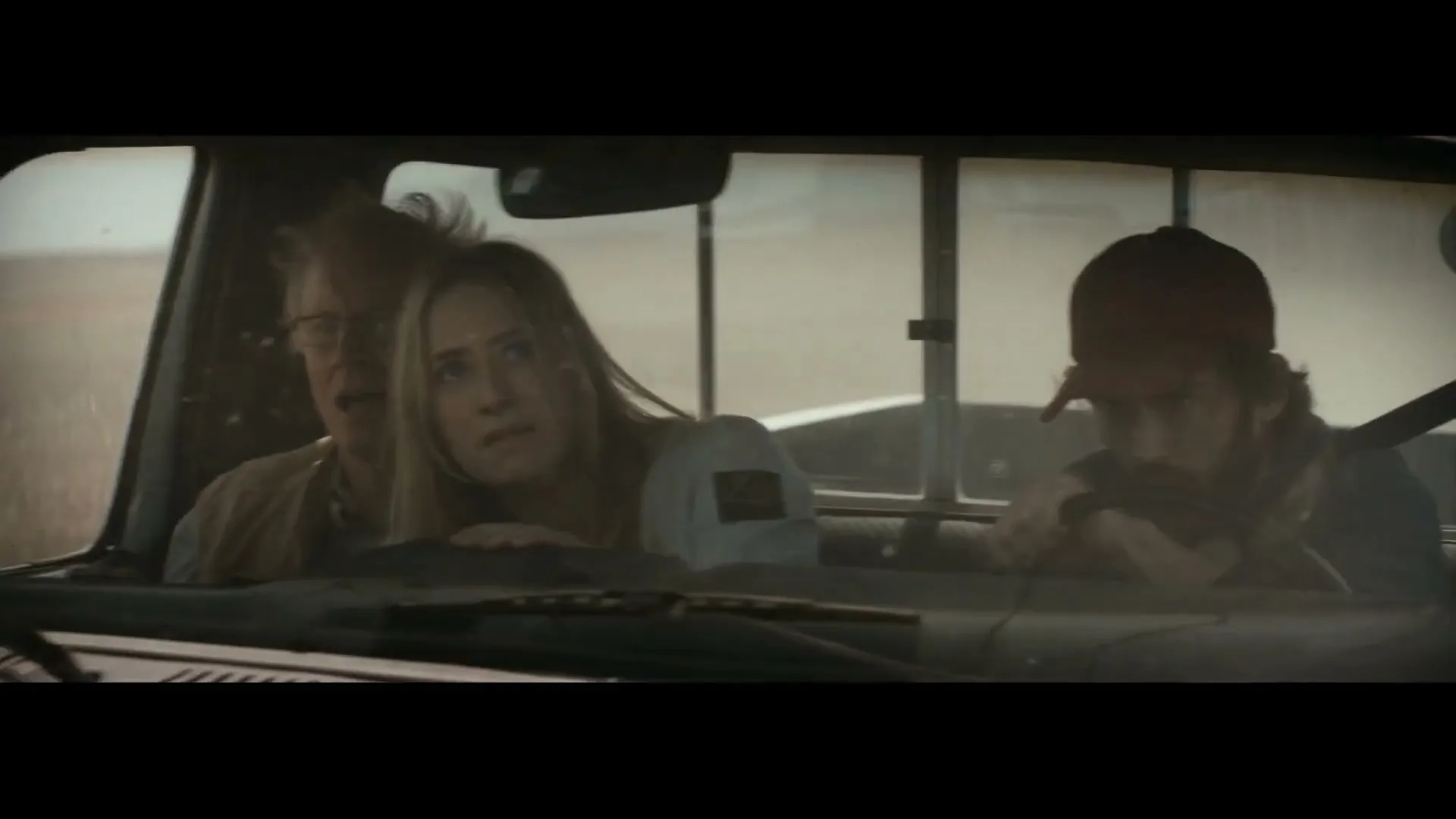 Three people, a man, a woman, and a younger man, are seen through the windshield of a moving pickup truck, looking scared as they drive through a dusty landscape.
