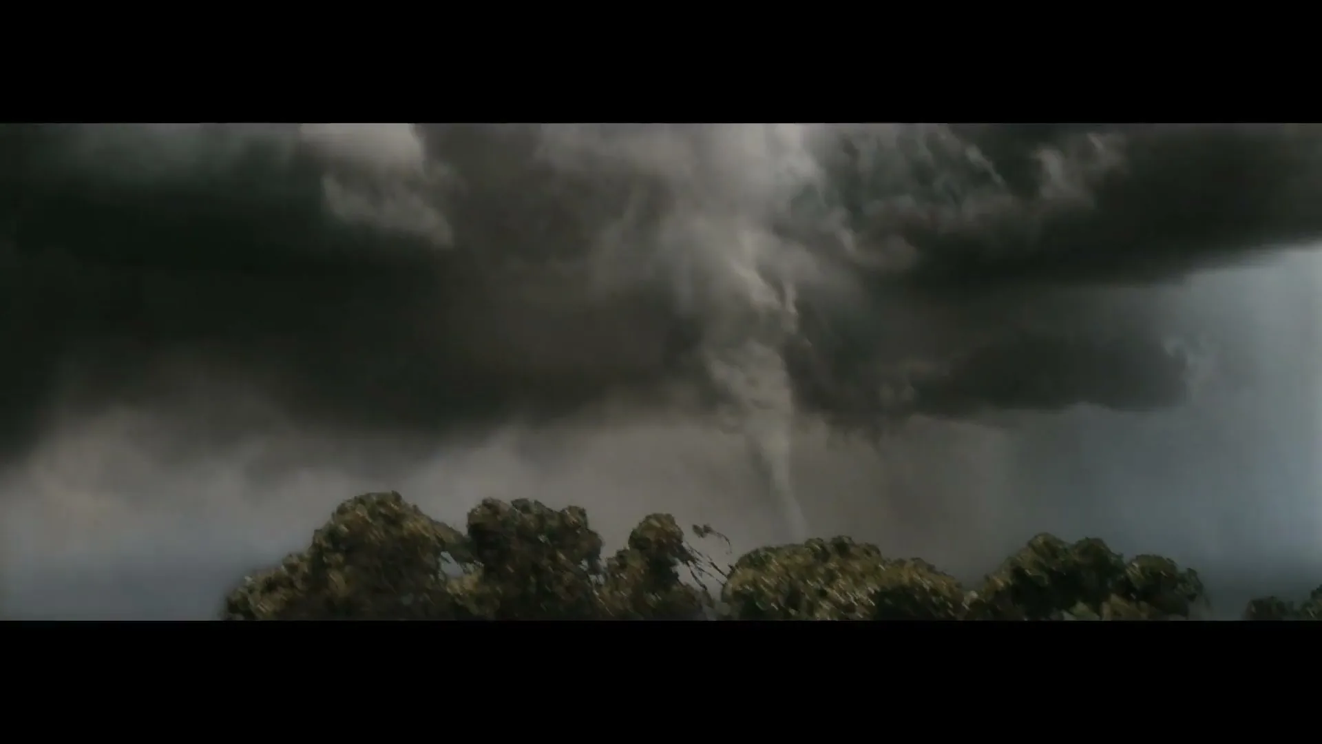 A powerful tornado descends from dark, stormy clouds, touching down behind a line of trees in a dramatic, wide shot.