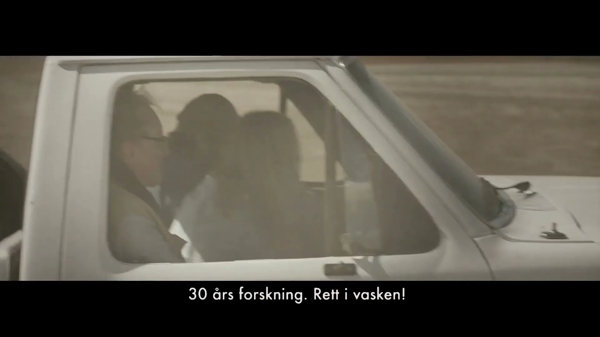 A man and a woman are seen inside a white pickup truck, driving through a dusty, barren landscape under a bright sky.
