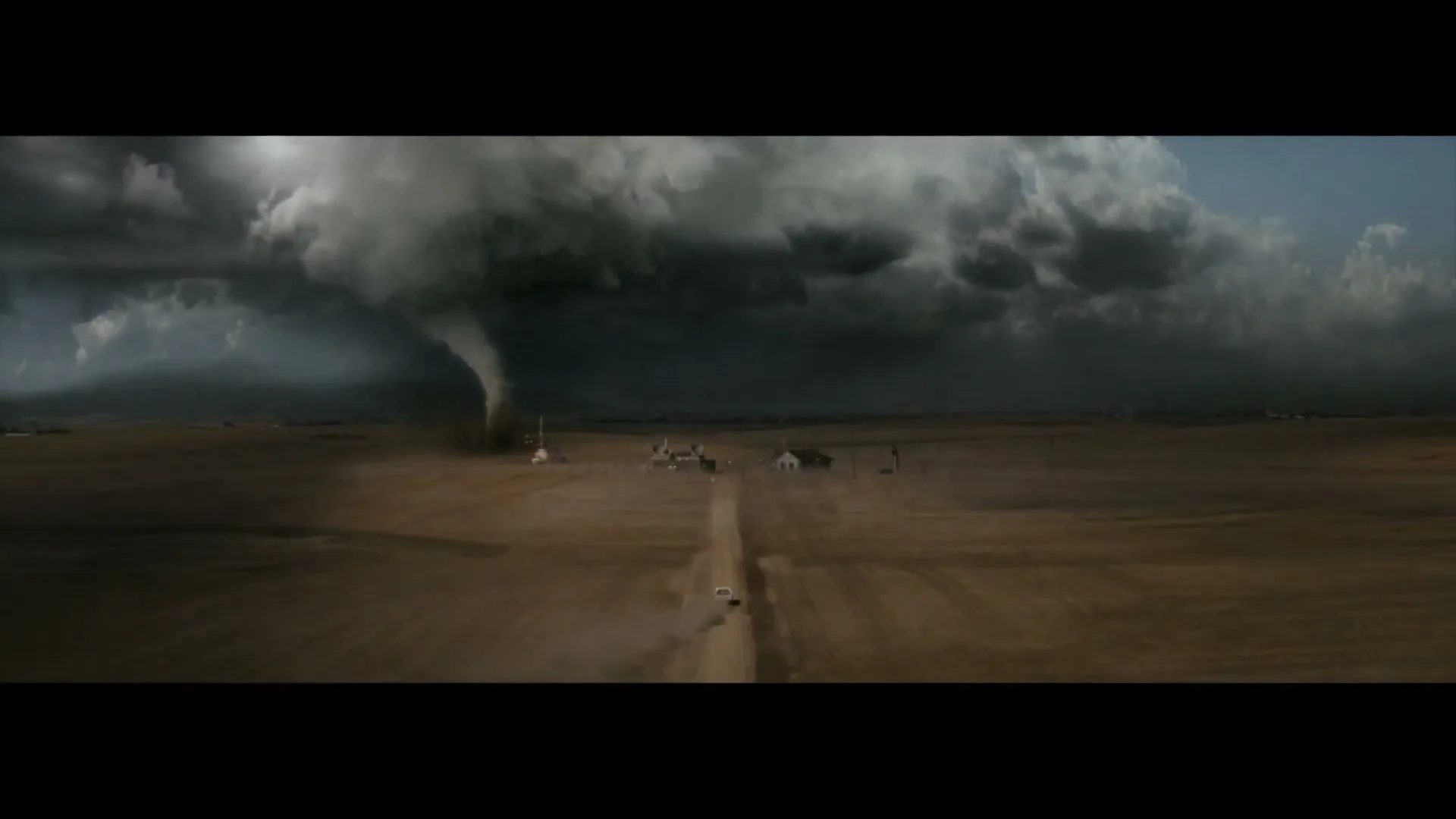 A wide shot shows a massive tornado looming over a small farm in a vast, dry field, with a car driving away on a dirt road.