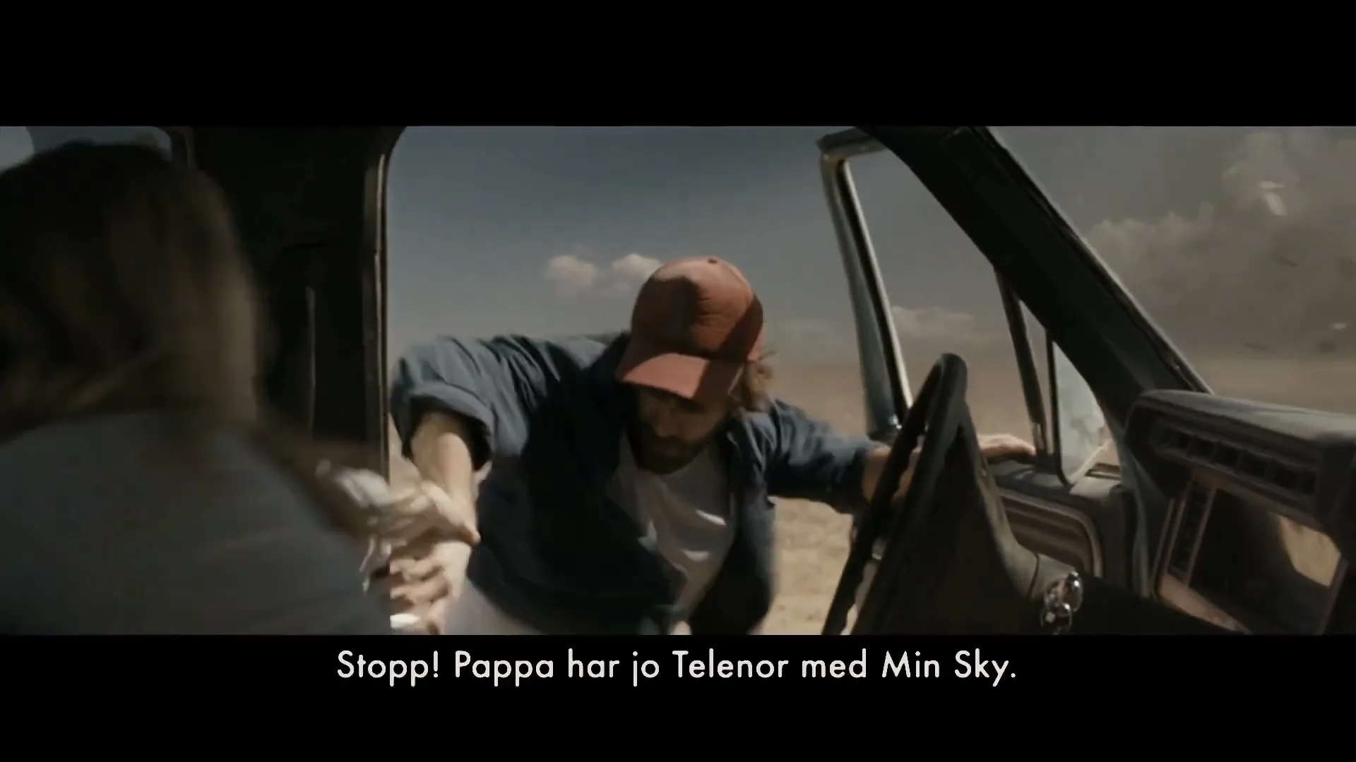 A man in a red cap and blue shirt leans out of a truck window, struggling against strong winds, while a woman tries to hold him back in a desert landscape.