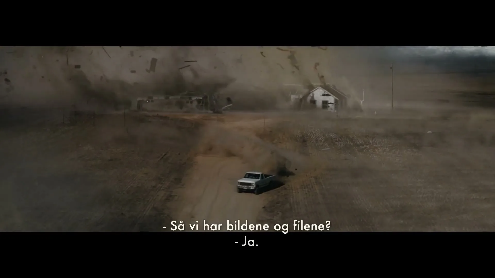 A white pickup truck drives away from a massive tornado destroying two houses in a desolate, dusty landscape under a dark sky.
