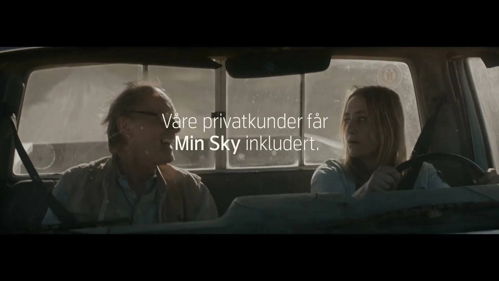 A man and a woman are sitting in a dusty pickup truck, looking forward and talking, with the man smiling.